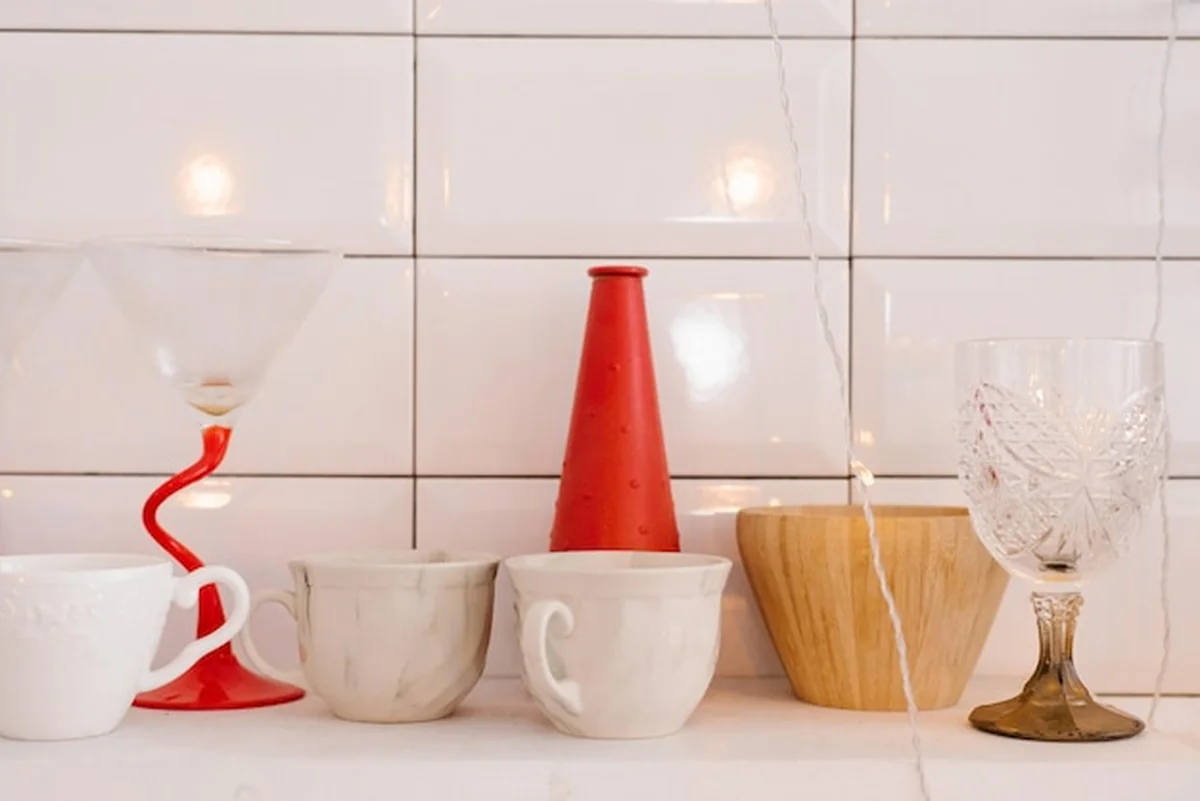 Białe porcelanowe lub ceramiczne kubki, drewniane naczynia, czerwony wazon i szklanka do wina na półce kuchennej